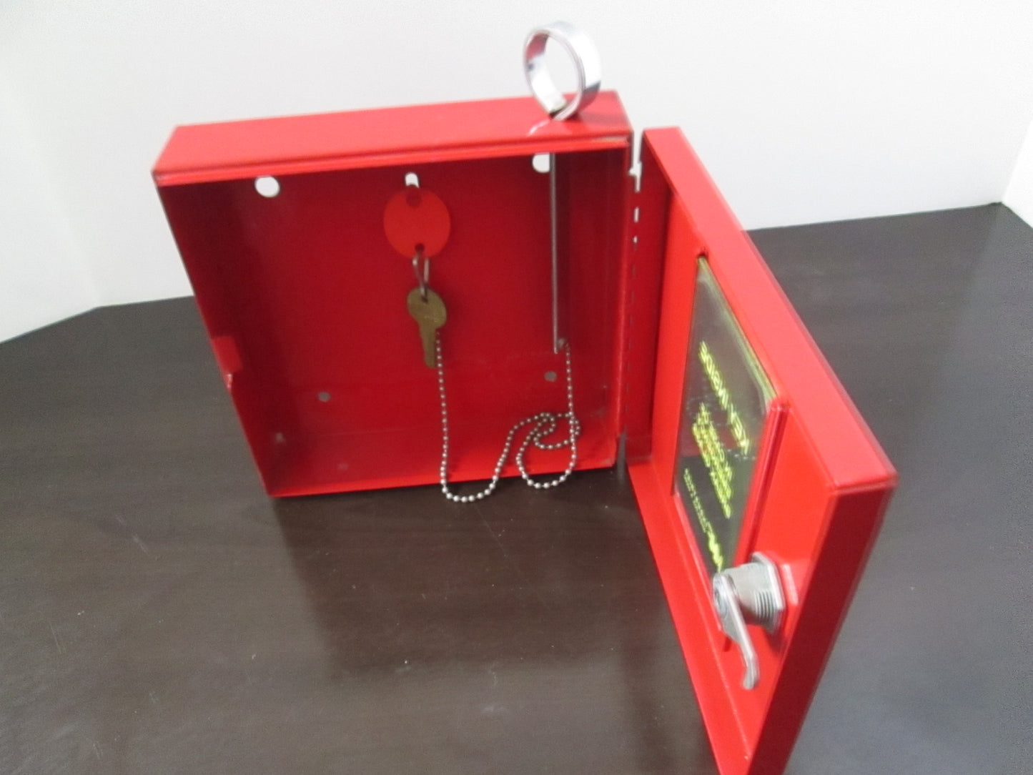 A bright red heavy gauge steel emergency key box with a lock on the front and a key inserted into the lock. The box is mounted on a table and has a digital display and a key lock.