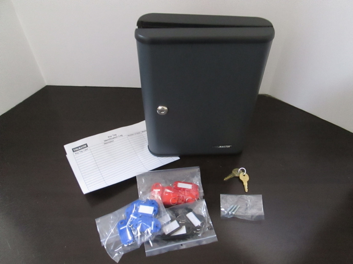 A gray metal key storage cabinet with hook-style key hangers, a cam lock on the door, and two keys. The cabinet is on a table with a paper with '30' on it, indicating its capacity to store 30 keys.